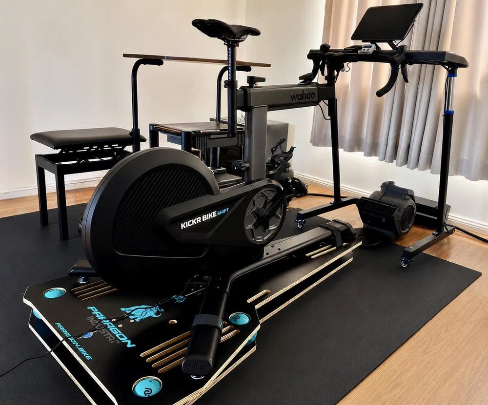 A home gym setup featuring a black indoor P3 Full-Motion Rocker plate for the Wahoo Kickr Bike Shift a black mat, photographed in a room with wood floors and curtains.